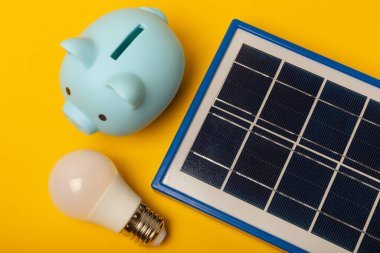 Güneş paneli, öncü lamba ve sarı arka planda domuz kumbarası olan düz bir kompozisyon. Tasarruf ve temiz enerji kavramı. Ekoloji ve sürdürülebilir kalkınma kavramı.