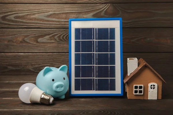 Flat lay composition with solar panel, led lamp, house model, money and piggy bank on brown wood. The concept of saving money and clean energy. Concept of ecology and sustainable development.