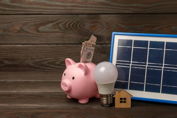 Flat lay composition with solar panel, led lamp, house model, money and piggy bank on brown wood. The concept of saving money and clean energy. Concept of ecology and sustainable development.