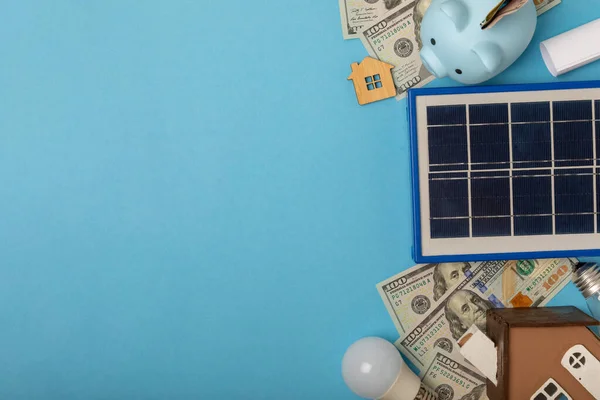 Flat lay composition with solar panel, led lamp, house model, money and piggy bank on blue background. The concept of saving money and clean energy. Concept of ecology and sustainable development.