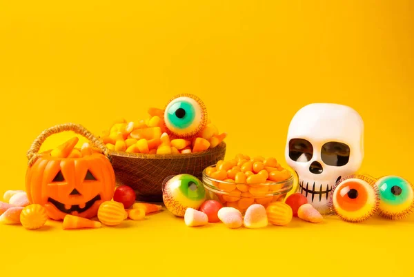 Halloween candy corn,jelly eye, pumpkin candy, sugar skull in different bowls on yellow background.Classic candy sweets for Halloween with.Halloween holiday concept with candy corn and Jack-o-lantern.
