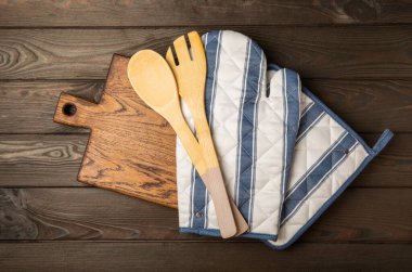 Ahşap mutfak aletleri, kesim tahtası, tutacağı ve eldiven masanın üstünde. Mutfak eldiveni ve koruyucu fırın eldivenleri masanın üstünde. Mutfak eşyaları. Mutfak aksesuarları. Yakın çekim. Üst manzara..