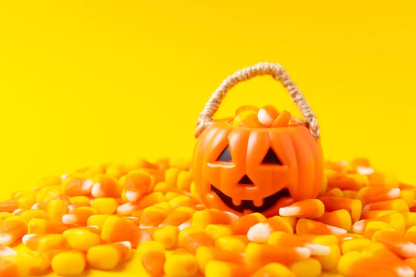 Halloween pumpkin Jack-o-lantern bucket with candy corn for celebration halloween. Classic white, orange and yellow Halloween lollipops. Sweet pumpkin gummies in a wooden bowl on a yellow background.