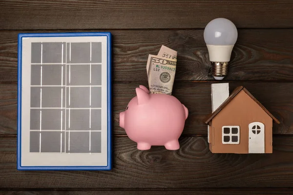 Flat lay composition with solar panel, led lamp. house model and a piggy bank on a brown table. The concept of saving money and clean energy. The concept of ecology and sustainable development.