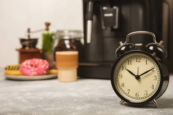コーヒーマシンの背景に目覚まし時計 台所のテーブルの上にある黒い目覚まし時計は アロマコーヒーとコーヒーマシンのカップを背景にしています 陽気な朝のコンセプト テキスト用の場所 コピーする場所 — ストック写真