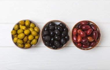 Beyaz ahşap zemin üzerinde yeşil, siyah ve kırmızı zeytinler. Taze zeytin yapraklı zeytin yağı ve kâse içinde çeşitli zeytin türleri. Uzayı kopyala Mesaj için yer. Akdeniz yemekleri. Vejetaryen.