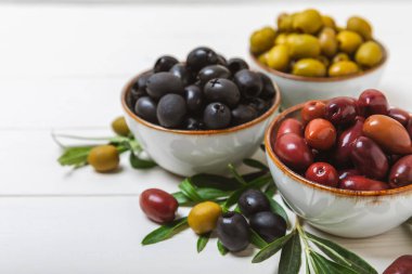 Beyaz ahşap zemin üzerinde yeşil, siyah ve kırmızı zeytinler. Taze zeytin yapraklı zeytin yağı ve kâse içinde çeşitli zeytin türleri. Uzayı kopyala Mesaj için yer. Akdeniz yemekleri. Vejetaryen.