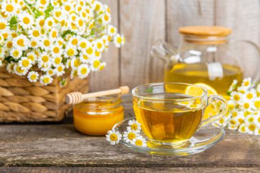 Kahverengi ahşap bir masada, içinde bal, limon ve papatya buketi olan cam bir fincanda papatya çayı. Yakın plan. Uzayı kopyala sağlıklı bitkisel içecekler, bağışıklık çayı. Doğal şifacı kavramı. Metin için yer..