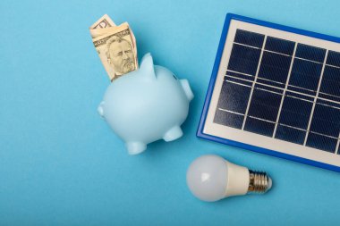 Güneş paneli, öncü lamba ve mavi arka planda domuz kumbarası olan düz bir kompozisyon. Tasarruf ve temiz enerji kavramı. Ekoloji ve sürdürülebilir kalkınma kavramı.