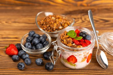 Kahverengi mutfak masasında yoğurt, taze badem, yabanmersini, ahududu, şeftali ve çilekli granola kasesi..