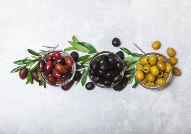 Açık mermer zemin üzerinde yeşil, kırmızı ve siyah zeytin seti. Kâselerde çeşitli zeytin ve taze zeytin yaprakları. Vejetaryen. Zeytin meyveleri. Mesaj için yer. Boşluğu kopyala.