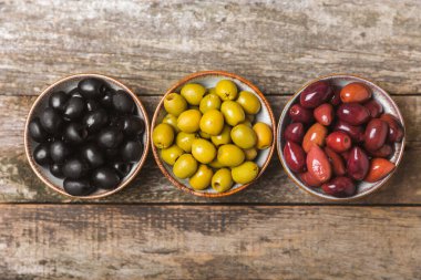 Yeşil, siyah ve kırmızı zeytinler, kahverengi ahşap arka planda zeytin yağı. Bir kâse taze zeytin ve taze zeytin yaprakları. Vejetaryen. Zeytin meyveleri. Mesaj için yer. Boşluğu kopyala.