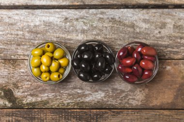 Yeşil, siyah ve kırmızı zeytinler, kahverengi ahşap arka planda zeytin yağı. Bir kâse taze zeytin ve taze zeytin yaprakları. Vejetaryen. Zeytin meyveleri. Mesaj için yer. Boşluğu kopyala.