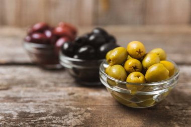 Yeşil, siyah ve kırmızı zeytinler, kahverengi ahşap arka planda zeytin yağı. Bir kâse taze zeytin ve taze zeytin yaprakları. Vejetaryen. Zeytin meyveleri. Mesaj için yer. Boşluğu kopyala.