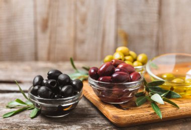 Yeşil, siyah ve kırmızı zeytinler, kahverengi ahşap arka planda zeytin yağı. Bir kâse taze zeytin ve taze zeytin yaprakları. Vejetaryen. Zeytin meyveleri. Mesaj için yer. Boşluğu kopyala.