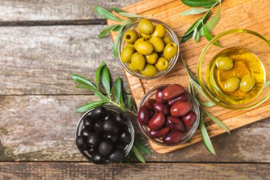 Yeşil, siyah ve kırmızı zeytinler, kahverengi ahşap arka planda zeytin yağı. Bir kâse taze zeytin ve taze zeytin yaprakları. Vejetaryen. Zeytin meyveleri. Mesaj için yer. Boşluğu kopyala.