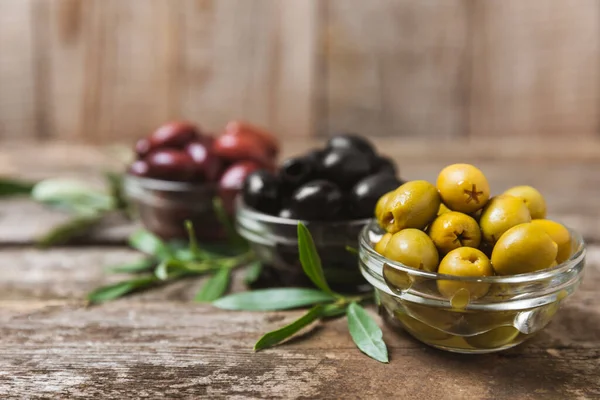 Yeşil, siyah ve kırmızı zeytinler, kahverengi ahşap arka planda zeytin yağı. Bir kâse taze zeytin ve taze zeytin yaprakları. Vejetaryen. Zeytin meyveleri. Mesaj için yer. Boşluğu kopyala.