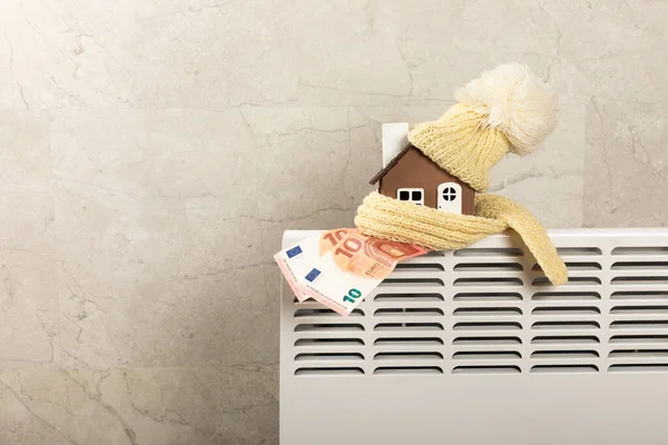 家庭取暖散热器 房子的模型 用围巾和帽子包裹在室内散热器上 靠在灰色的墙壁上 冬季取暖的效率 加热季节的概念 文本空间 复制空间 — 图库照片