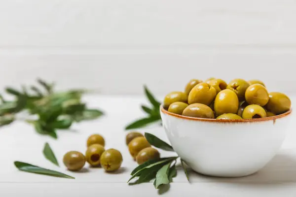 Greens olives on a white wooden background. Various types of olives in bowls and olive oil with fresh olive leaves. Copy space. Place for text. Mediterranean food. Vegan.
