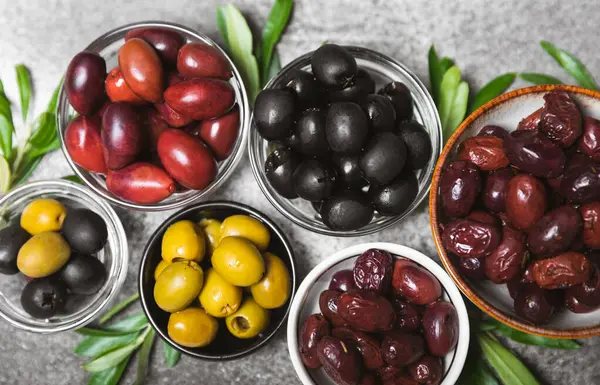 A set of green, red and black olives on a marble background. Various types of olives in bowls and fresh olive leaves. Vegan. Olive fruits. Place for text. Copy space.