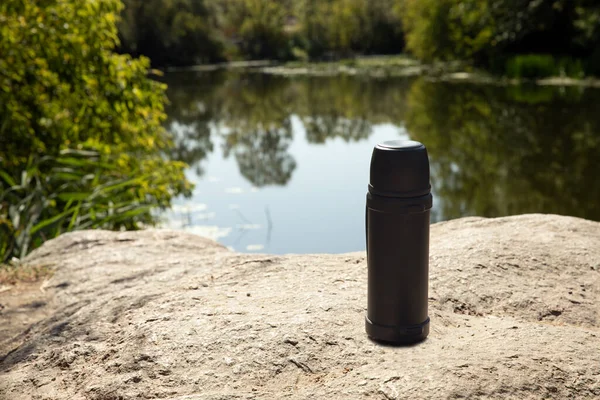 Thermos with delicious hot tea or coffee on the mountain. Autumn forest background is blurred. Warming drink in a thermos against the backdrop of a lake in the forest. Thematic tourism and travel