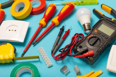 Mavi arka planda elektrikli aletler. Multimetre, inşaat bandı, elektrik bandı, tornavida, pense, otomatik yalıtım striptizcisi, soket ve LED lamba..