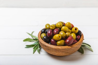 Beyaz ahşap zemin üzerinde yeşil, kırmızı ve siyah zeytin seti. Tahta kaselerde çeşitli zeytinler ve taze zeytin yaprakları. Uzayı kopyala Mesaj için yer. Düz lei. Lezzetli.
