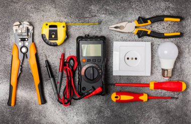 Arka planda elektrikli aletler. Multimetre, inşaat bandı, elektrik bandı, tornavida, pense, otomatik yalıtım striptizcisi, soket ve LED lamba..