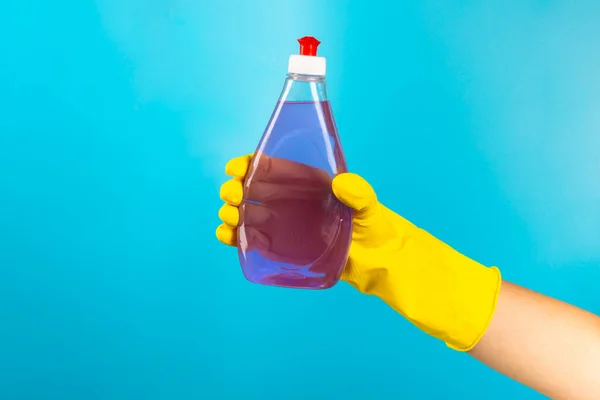 A cleaner's hand in a yellow rubber protective glove holding a bottle with a cleaning chemical on a blue background. Commercial cleaning company. Spring regular cleaning. Space for text or logo.