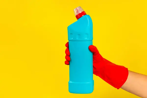A cleaner's hand in a red rubber protective glove holding a bottle with a cleaning chemical on a yellow background. Commercial cleaning company. Spring regular cleaning. Space for text or logo.