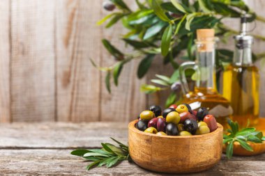 Yeşil, siyah ve kırmızı zeytinler, kahverengi ahşap arka planda zeytin yağı. Bir kâse taze zeytin ve taze zeytin yaprakları. Vejetaryen. Zeytin meyveleri. Mesaj için yer. Boşluğu kopyala.