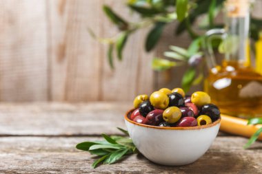 Yeşil, siyah ve kırmızı zeytinler, kahverengi ahşap arka planda zeytin yağı. Bir kâse taze zeytin ve taze zeytin yaprakları. Vejetaryen. Zeytin meyveleri. Mesaj için yer. Boşluğu kopyala.