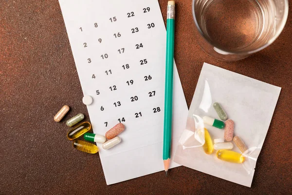 Vitamins and supplements. Variety of vitamin tablets with a glass of water and a calendar on a brown background. Multivitamins for every day. Nutritional supplements. Flat lei.Time to drink pills.