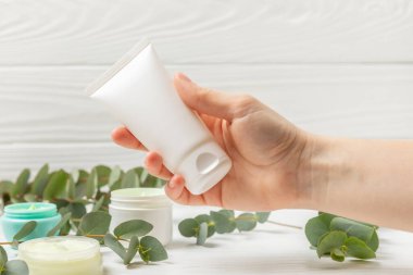 Kahverengi ahşap arka planda okaliptüs yaprakları olan kadınların ellerinde yüz, eller ve vücut için bir kavanoz nemlendirici kozmetik krem. Doğal organik ürün. Güzellik ve kaplıca konsepti. Vücut bakımı. Boşluğu kopyala.