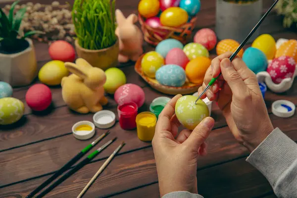 Woman paints Easter eggs at the kitchen wooden table.Happy Easter celebration concept.Colorful Easter eggs with different patterns.Paints,decorations for coloring eggs for holiday.Creative background.