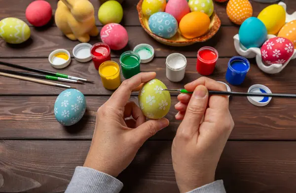 Woman paints Easter eggs at the kitchen wooden table.Happy Easter celebration concept.Colorful Easter eggs with different patterns.Paints,decorations for coloring eggs for holiday.Creative background.
