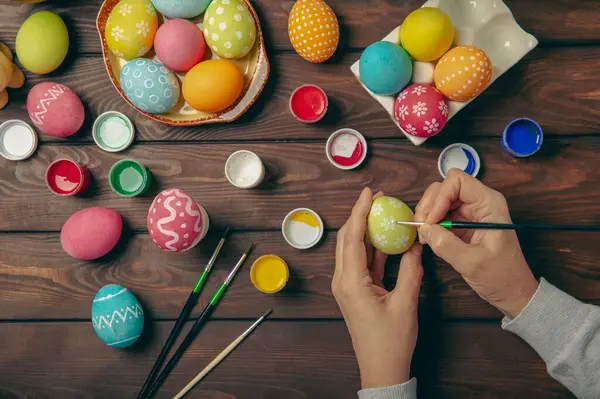 Woman paints Easter eggs at the kitchen wooden table.Happy Easter celebration concept.Colorful Easter eggs with different patterns.Paints,decorations for coloring eggs for holiday.Creative background.
