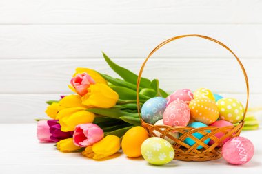 Renkli yumurtalarla dolu Paskalya sepeti ve ahşap bir masanın üzerinde bir buket lale. Paskalya kutlaması. El yapımı renkli Paskalya yumurtaları. Mesaj için yer. Boşluğu kopyala