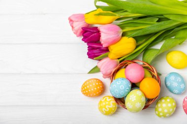 Renkli yumurtalarla dolu Paskalya sepeti ve ahşap bir masanın üzerinde bir buket lale. Paskalya kutlaması. El yapımı renkli Paskalya yumurtaları. Mesaj için yer. Boşluğu kopyala