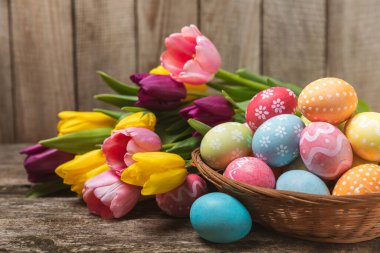 Renkli yumurtalarla dolu Paskalya sepeti ve ahşap bir masanın üzerinde bir buket lale. Paskalya kutlaması. El yapımı renkli Paskalya yumurtaları. Mesaj için yer. Boşluğu kopyala