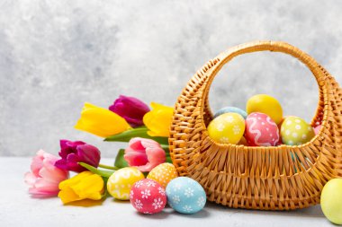Renkli yumurtalarla dolu Paskalya sepeti ve ahşap bir masanın üzerinde bir buket lale. Paskalya kutlaması. El yapımı renkli Paskalya yumurtaları. Mesaj için yer. Boşluğu kopyala