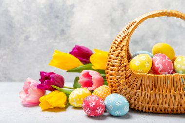 Renkli yumurtalarla dolu Paskalya sepeti ve ahşap bir masanın üzerinde bir buket lale. Paskalya kutlaması. El yapımı renkli Paskalya yumurtaları. Mesaj için yer. Boşluğu kopyala