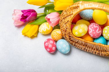 Renkli yumurtalarla dolu Paskalya sepeti ve ahşap bir masanın üzerinde bir buket lale. Paskalya kutlaması. El yapımı renkli Paskalya yumurtaları. Mesaj için yer. Boşluğu kopyala