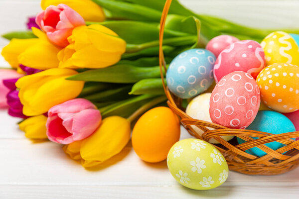 Easter basket filled with colorful eggs and a bouquet of tulips on a textured wooden table. Easter celebration concept. Colorful easter handmade decorated Easter eggs. Place for text. Copy space