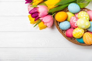 Renkli yumurtalarla dolu Paskalya sepeti ve ahşap bir masanın üzerinde bir buket lale. Paskalya kutlaması. El yapımı renkli Paskalya yumurtaları. Mesaj için yer. Boşluğu kopyala