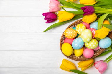 Renkli yumurtalarla dolu Paskalya sepeti ve ahşap bir masanın üzerinde bir buket lale. Paskalya kutlaması. El yapımı renkli Paskalya yumurtaları. Mesaj için yer. Boşluğu kopyala