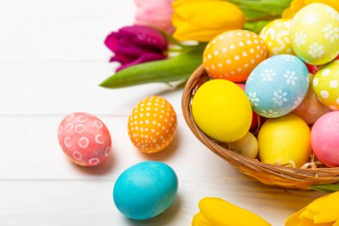 Renkli yumurtalarla dolu Paskalya sepeti ve ahşap bir masanın üzerinde bir buket lale. Paskalya kutlaması. El yapımı renkli Paskalya yumurtaları. Mesaj için yer. Boşluğu kopyala