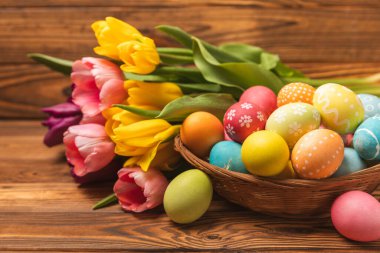 Renkli yumurtalarla dolu Paskalya sepeti ve ahşap bir masanın üzerinde bir buket lale. Paskalya kutlaması. El yapımı renkli Paskalya yumurtaları. Mesaj için yer. Boşluğu kopyala