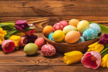 Renkli yumurtalarla dolu Paskalya sepeti ve ahşap bir masanın üzerinde bir buket lale. Paskalya kutlaması. El yapımı renkli Paskalya yumurtaları. Mesaj için yer. Boşluğu kopyala