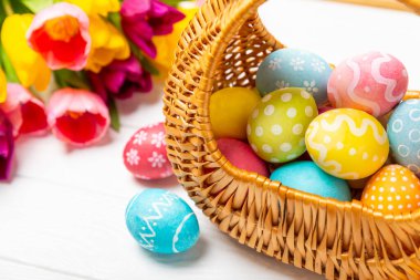 Renkli yumurtalarla dolu Paskalya sepeti ve ahşap bir masanın üzerinde bir buket lale. Paskalya kutlaması. El yapımı renkli Paskalya yumurtaları. Mesaj için yer. Boşluğu kopyala